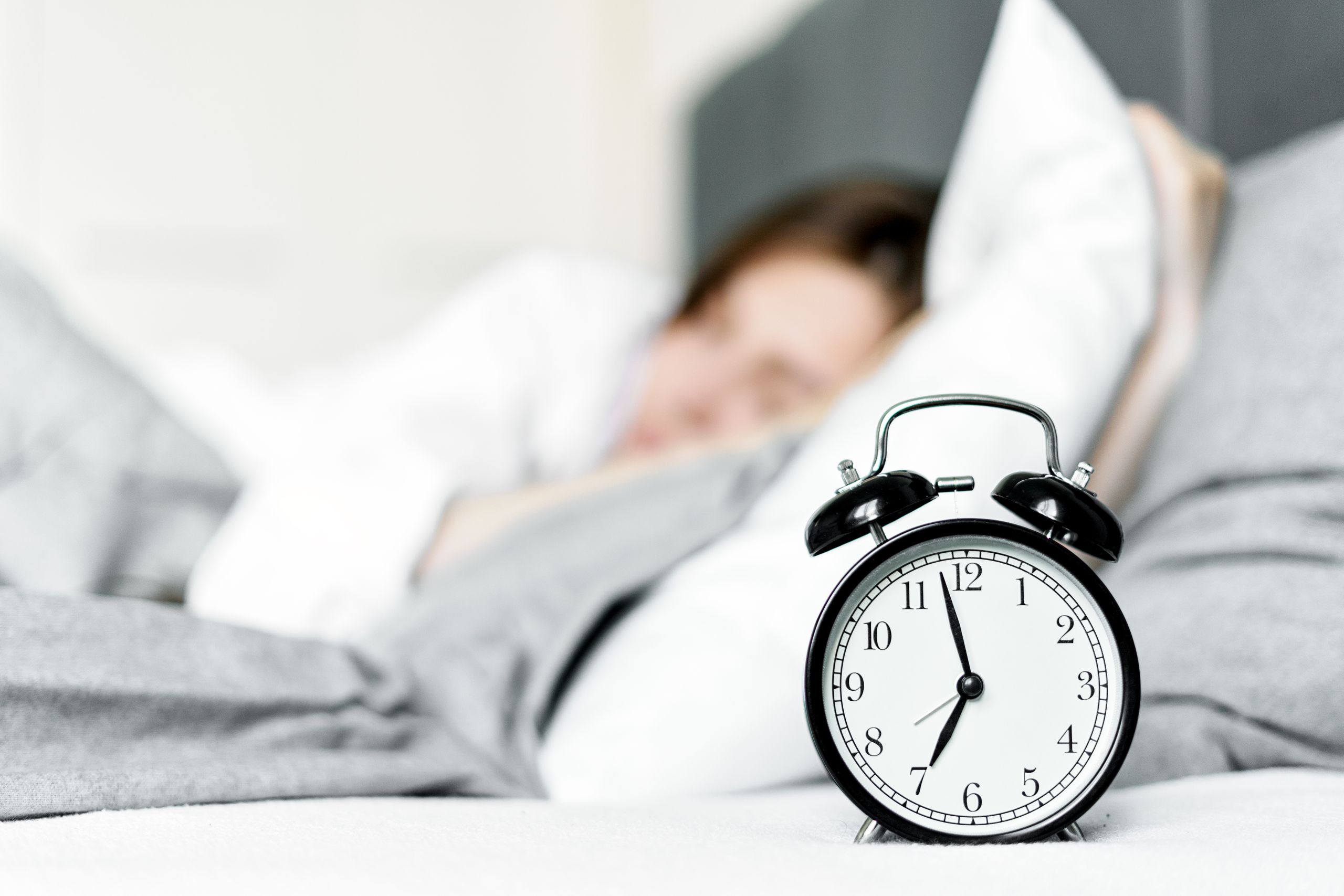 A retro alarm clock sits by a sleeping woman in a cozy bedroom setting.